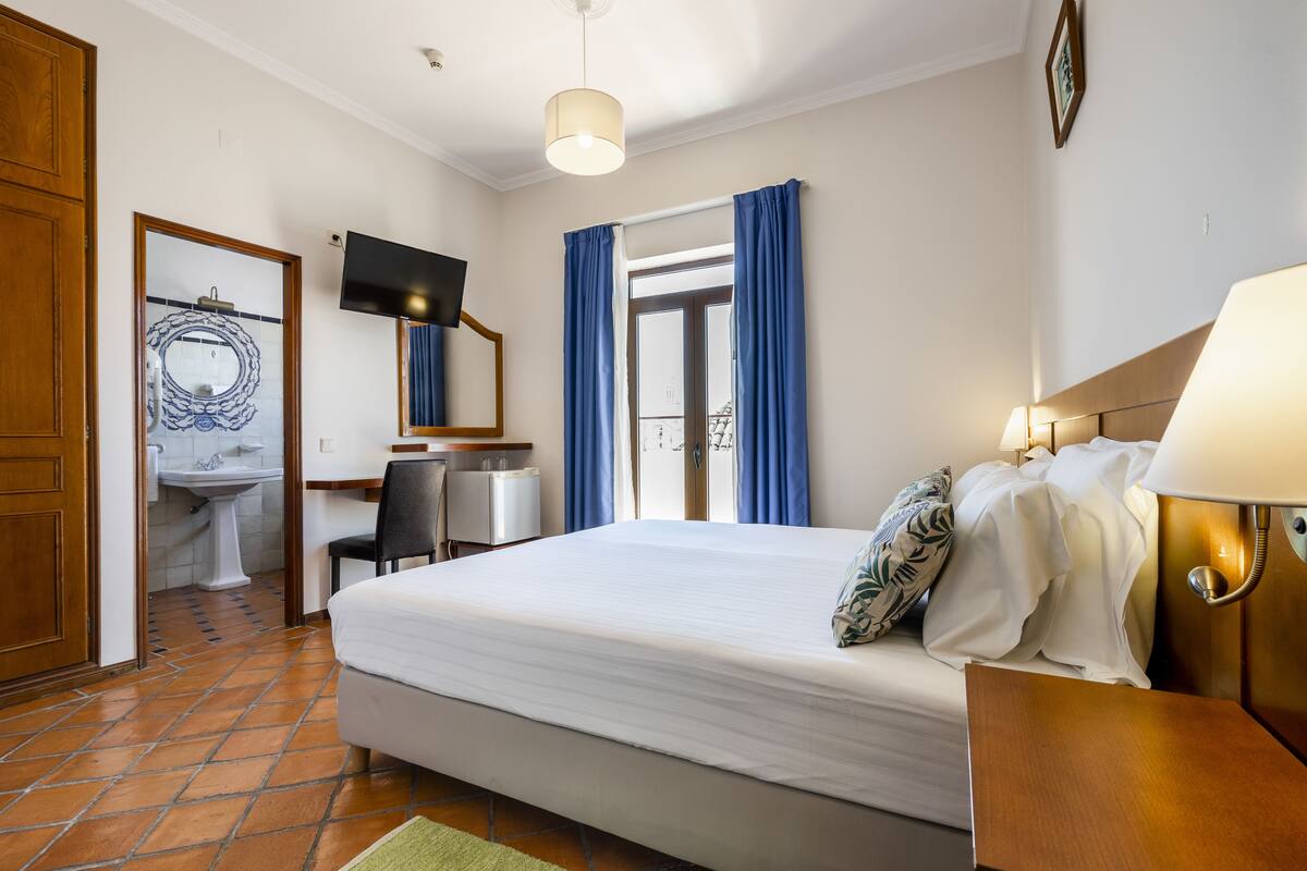 A hotel room with a bed, a desk, a chair, a TV, a bathroom with a sink and mirror, and a window with curtains.