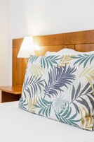 Una cama con sábana blanca y almohada con estampado de hojas tropicales.