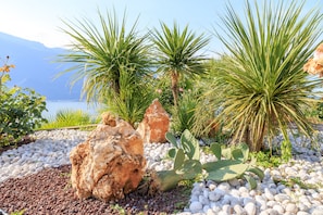 A garden with various plants and rocks, a body of water in the background, and mountains in the distance. - Hotel Cristina (Limone sul Garda)