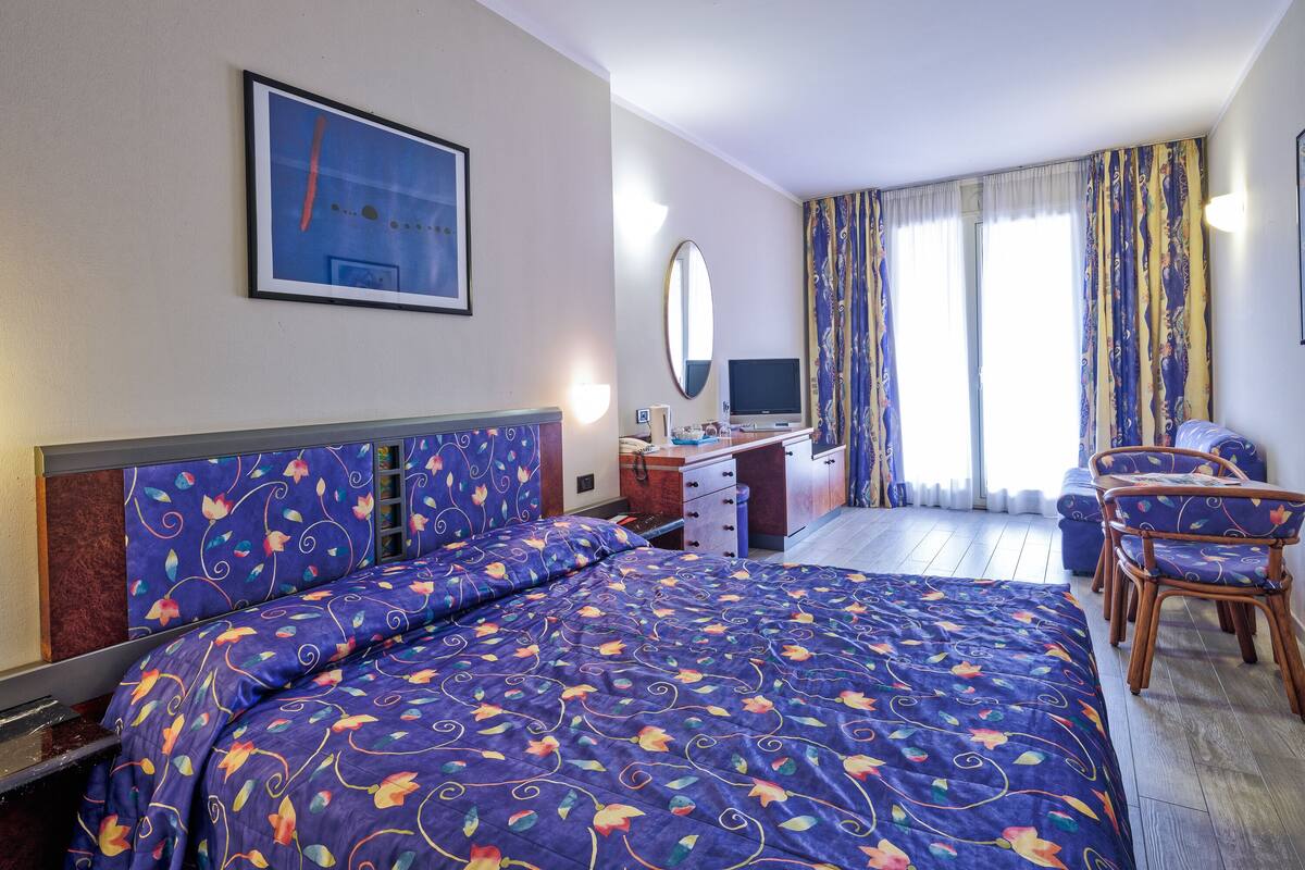 A hotel room with a large bed, a desk with a computer, a chair, and a window with curtains.
