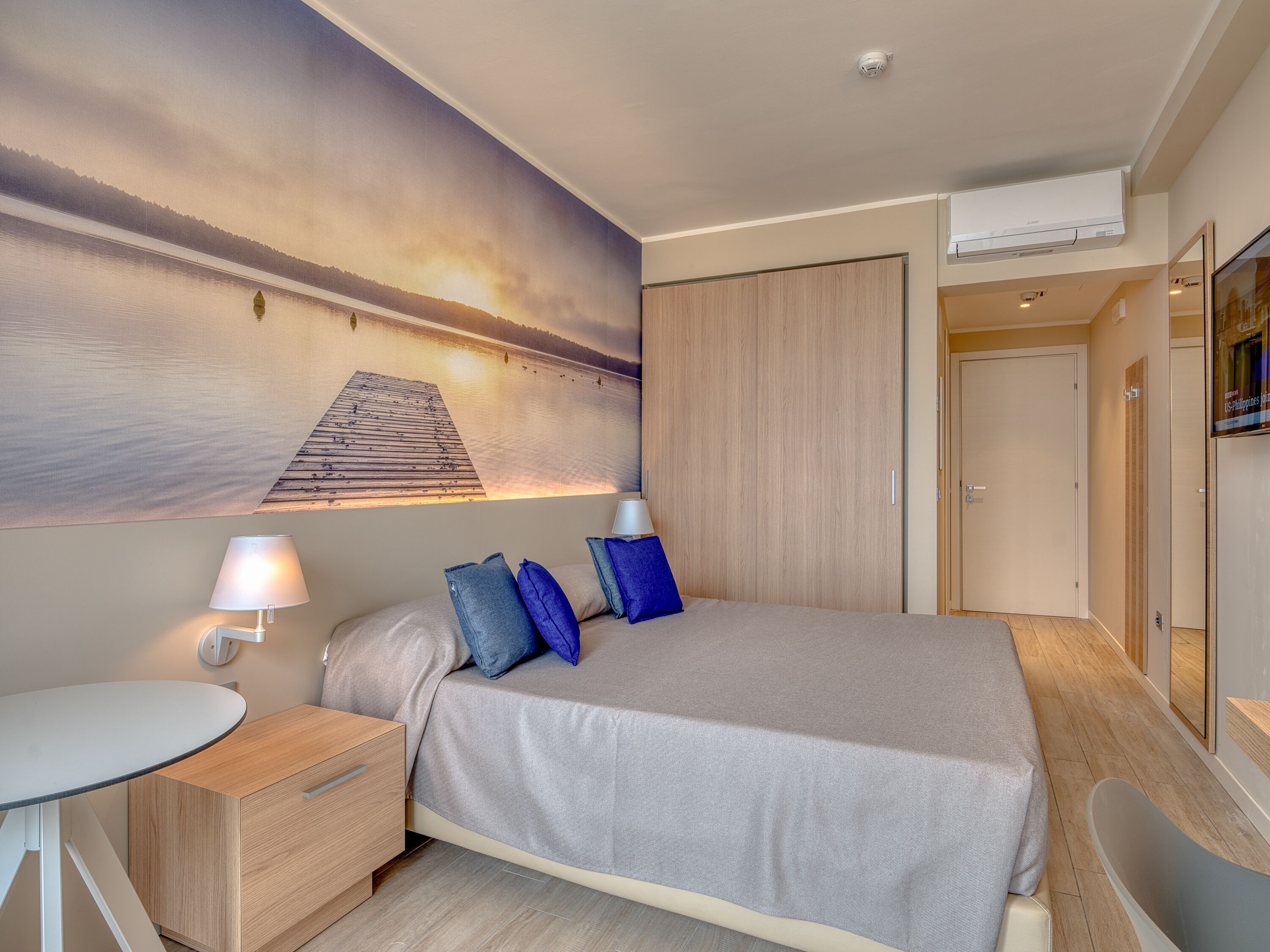 Un dormitorio con una cama, una mesita de noche, un escritorio, una lámpara de pared y un ventanal con vistas a un muelle y al agua.