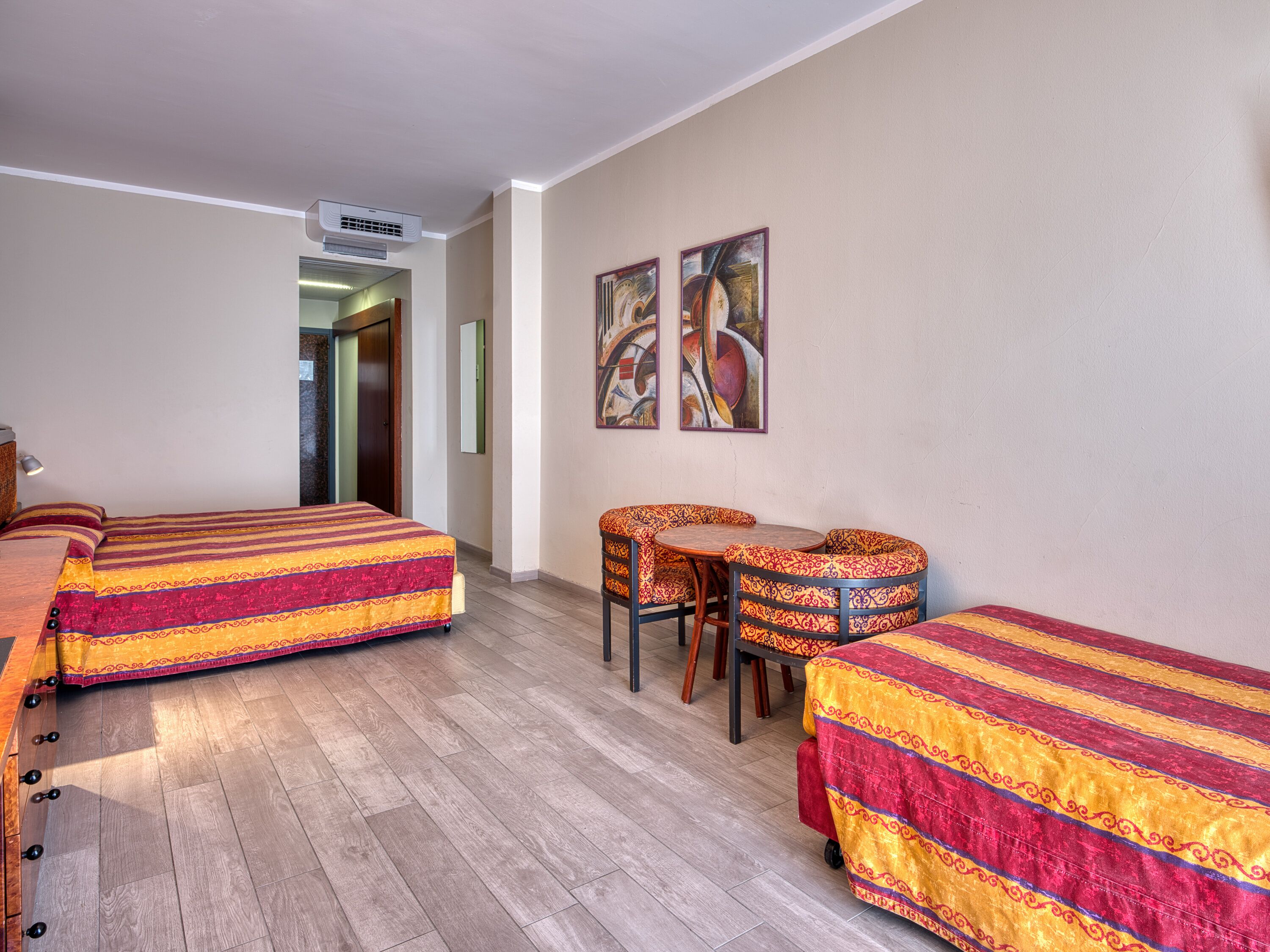 Habitación de hotel con dos camas, una mesita y sillas. Se observa una unidad de aire acondicionado en la pared y una puerta que da a otra habitación.
