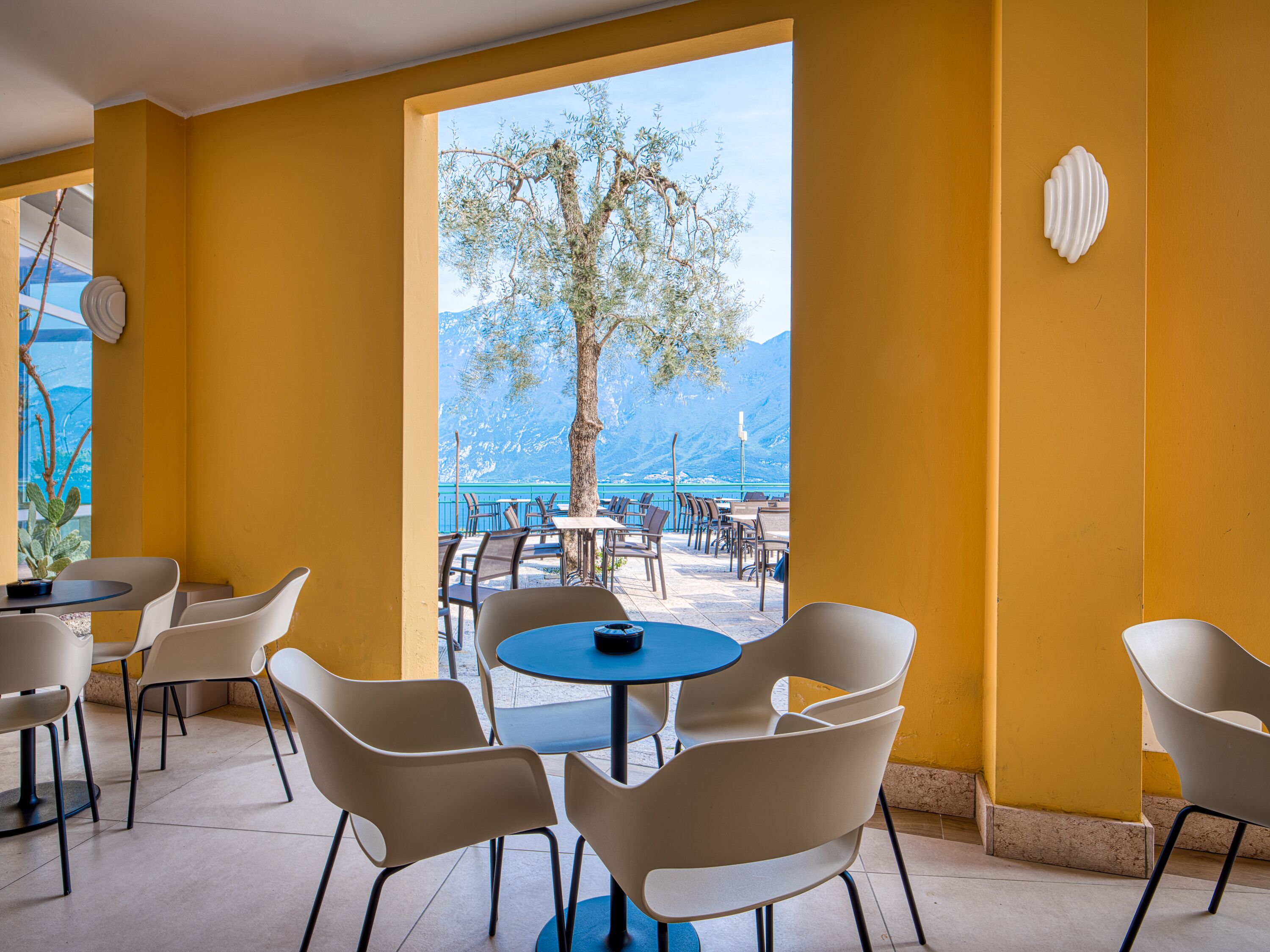 Restaurante com paredes amarelas, cadeiras e mesas brancas. Vista para uma árvore e montanhas através de grandes janelas.