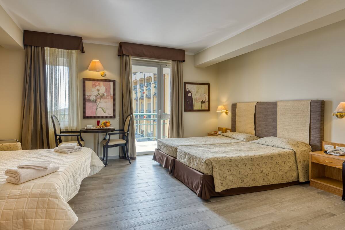 A hotel room with two beds, a desk, and a chair. There are two paintings on the walls, a window with curtains, and a balcony visible through the door.