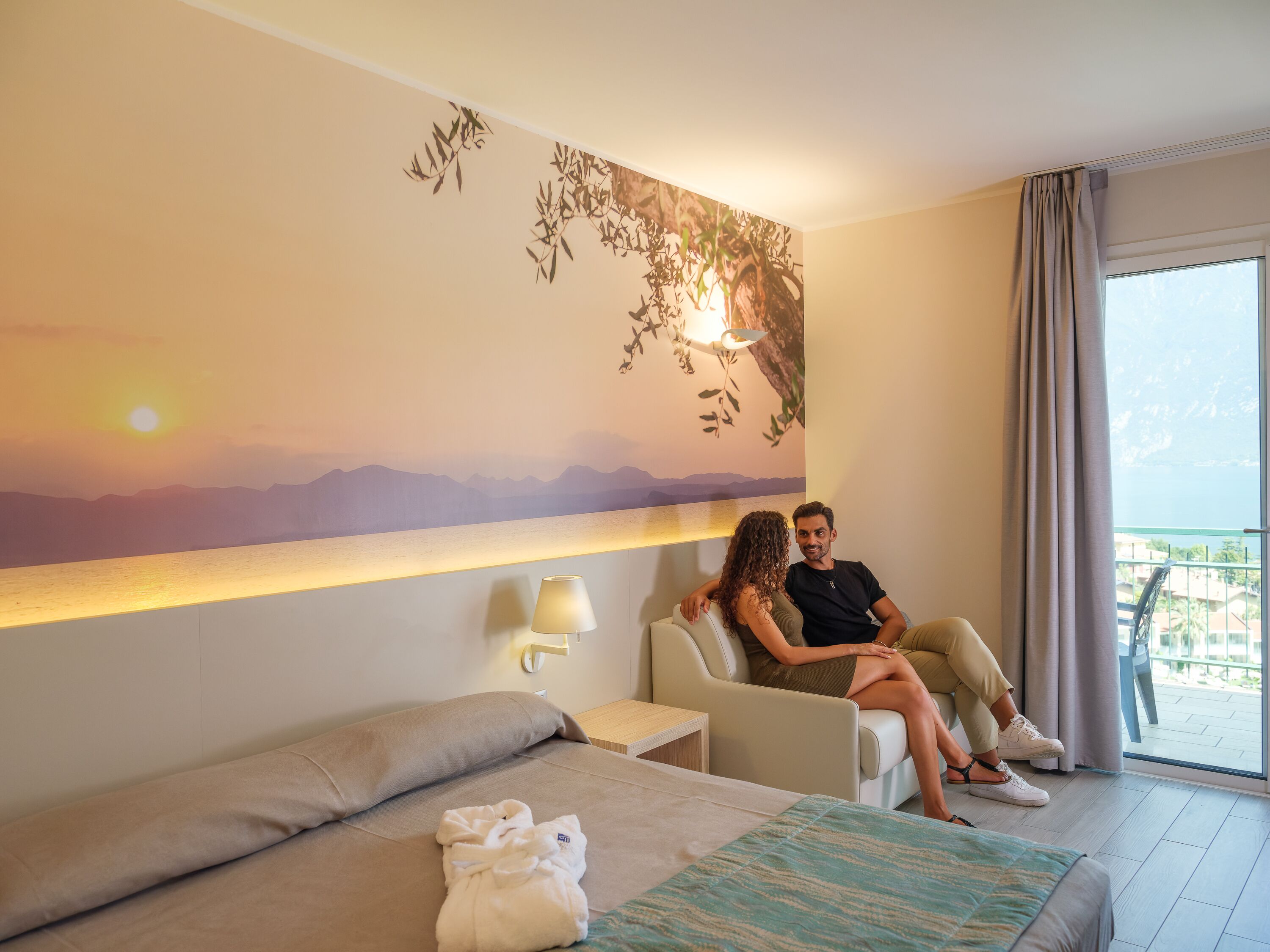 A hotel room with a bed, a sofa, a wall-mounted lamp, and a view of the outdoors.