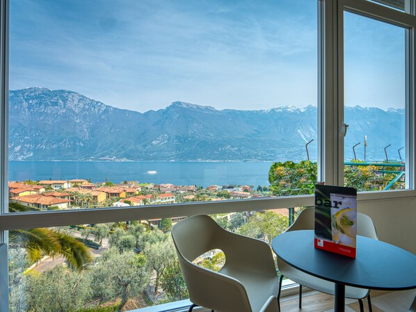 A room with a large window offering a view of a lake and mountains. There is a table with a chair and a menu on it. - Hotel Cristina (Limone sul Garda)