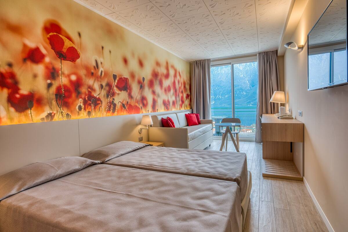 A modern hotel room with two beds, a sofa, a desk, and a large window with a view of the sea.