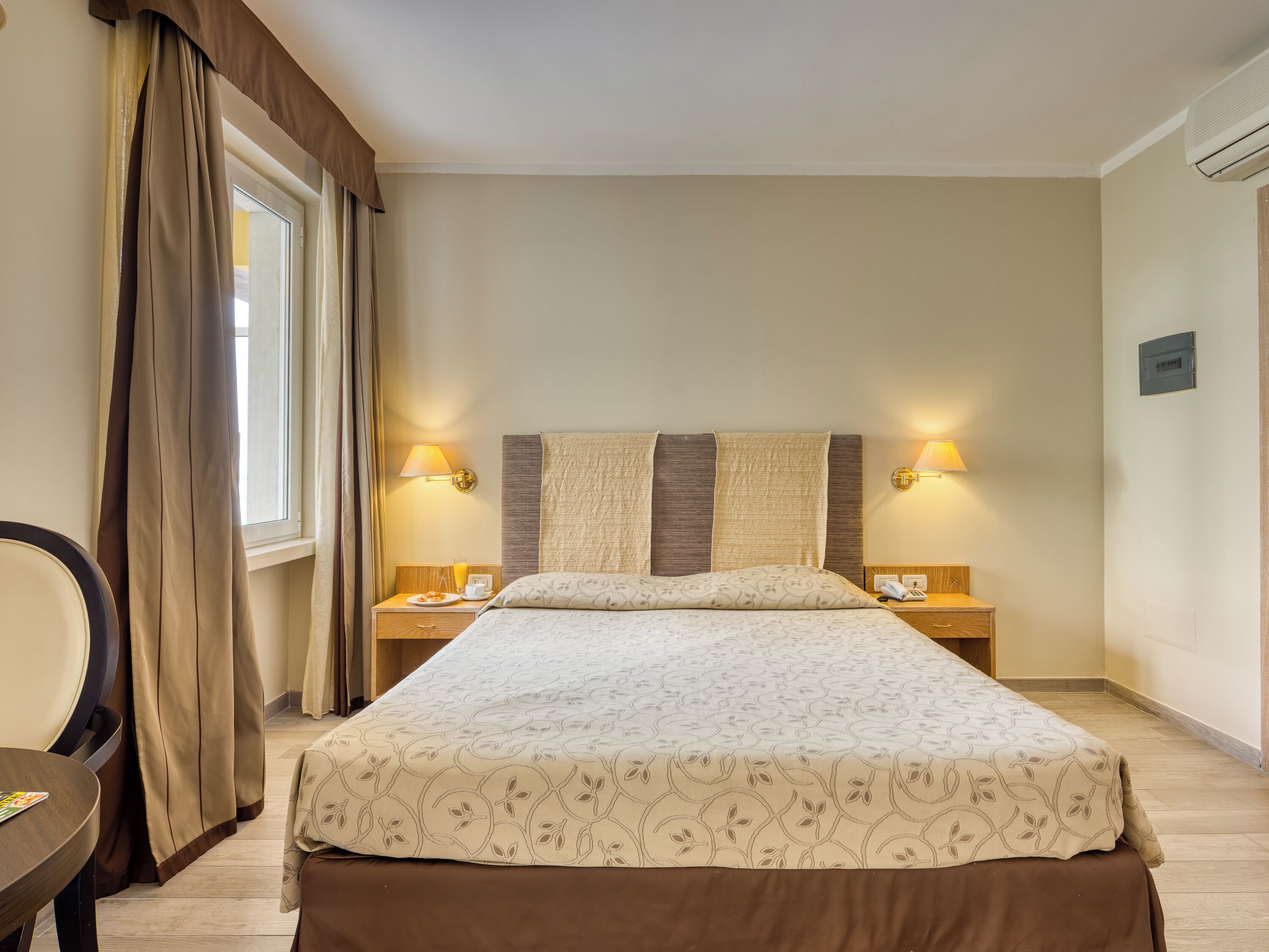 Una habitación de hotel con una cama grande, dos mesitas de noche, una silla y una ventana con cortinas.