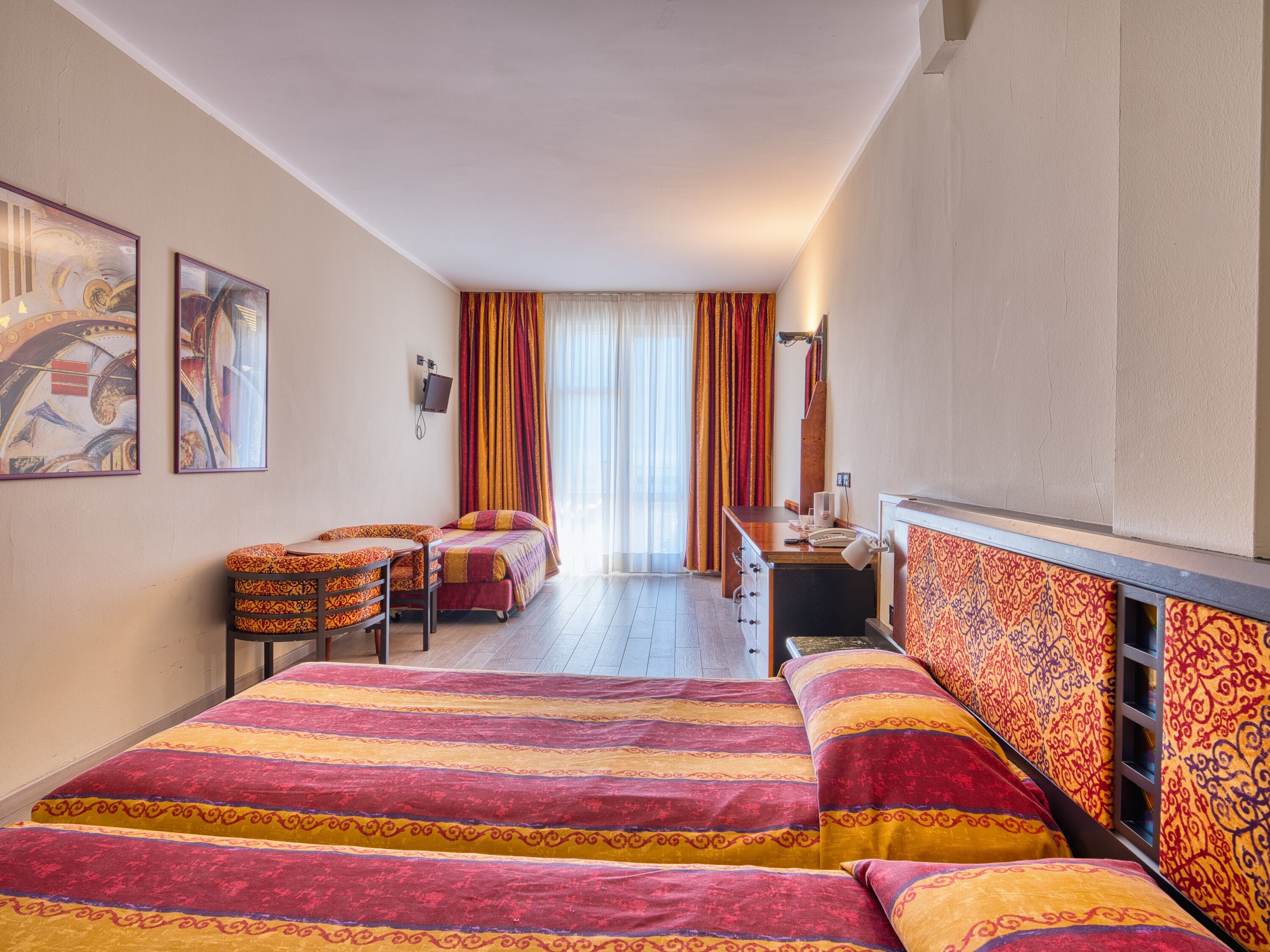 Ein Hotelzimmer mit Bett, Schreibtisch, Stuhl und einem kleinen Tisch. Vorhänge und ein Fenster sind vorhanden.
