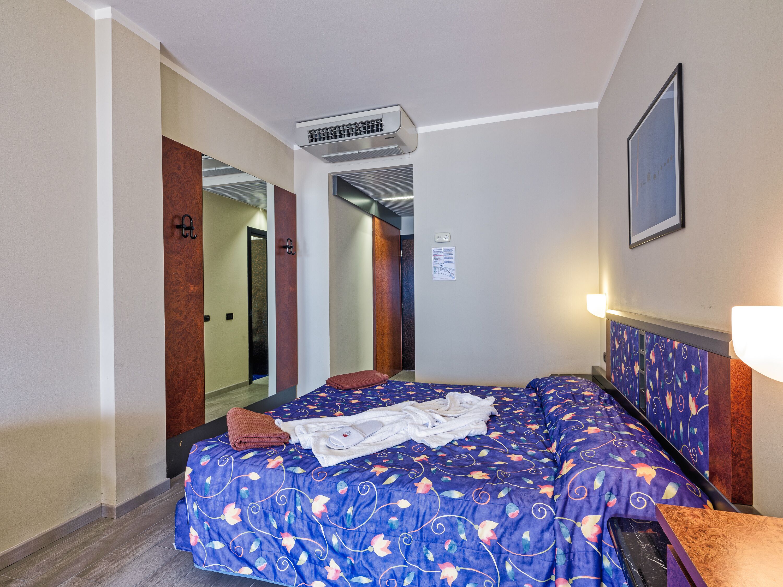 A hotel room with a large bed, an air conditioning unit, a mirror, and a framed picture on the wall.