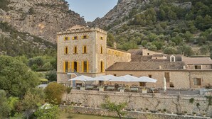Un bâtiment en pierre avec des volets jaunes, des tables à l’extérieur, protégées par des parasols, et une chaîne de montagnes en arrière-plan.