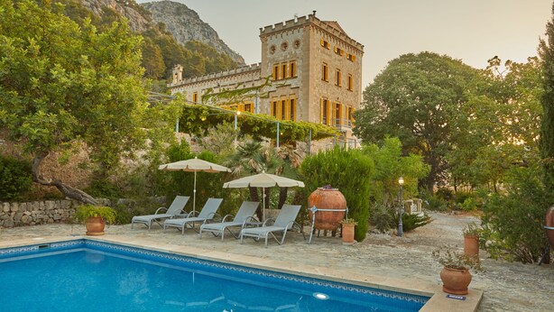 Un espace piscine avec des chaises longues et des parasols, un bâtiment en pierre doté d’une tour, et une végétation luxuriante.