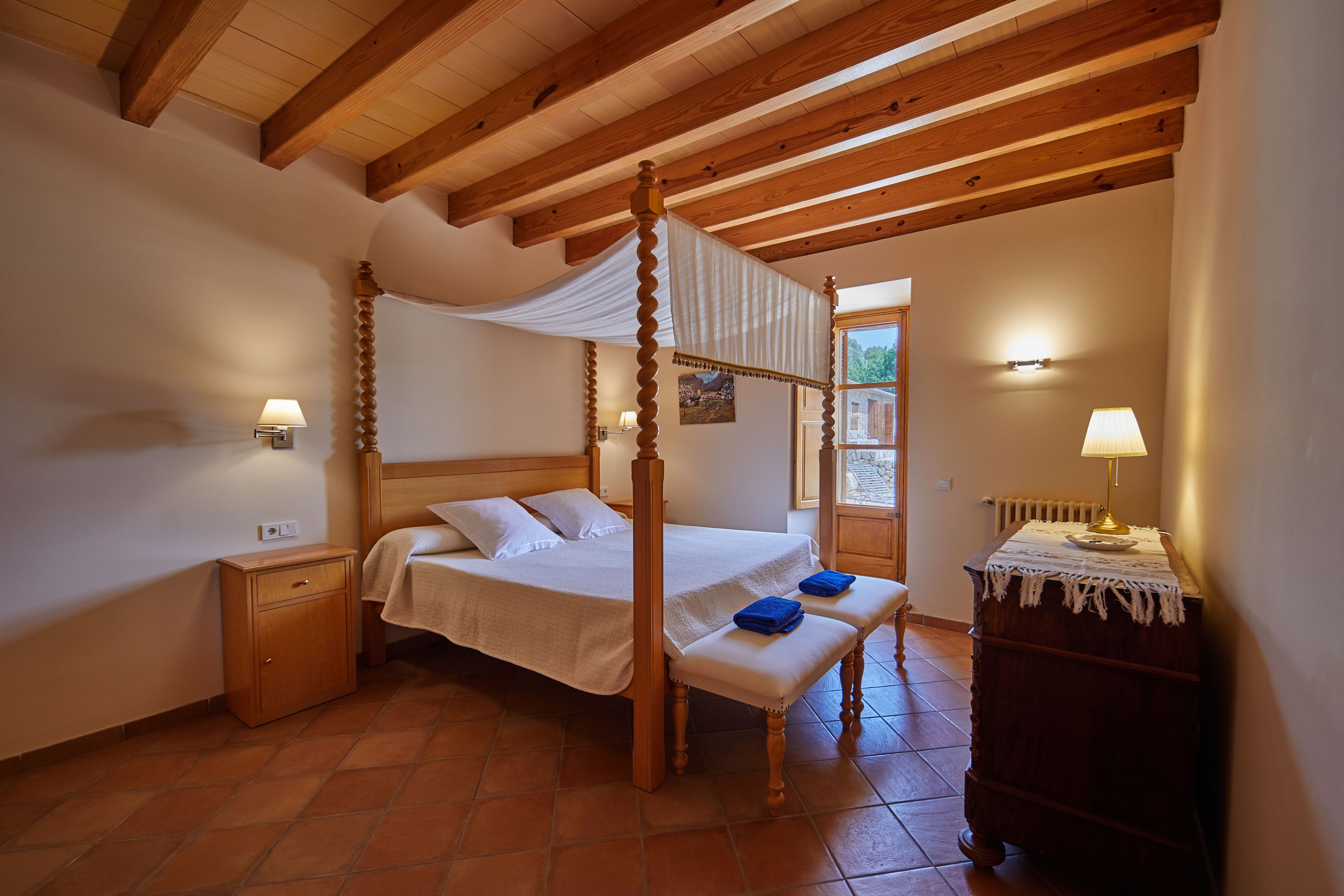 A four-poster bed with a canopy, a wooden nightstand, a bench, and a dresser with a lamp.