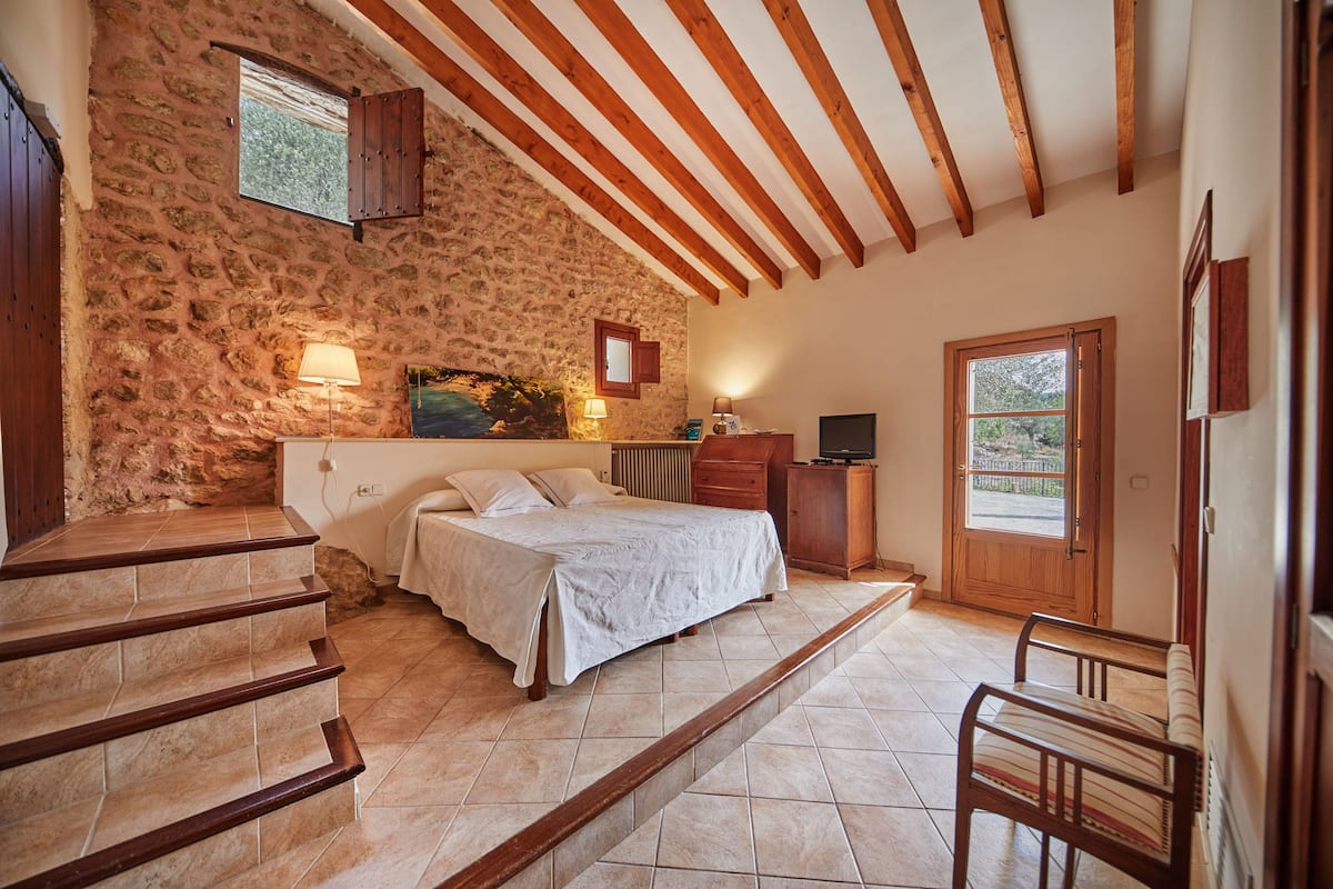 Ein Schlafzimmer mit Steinwand, Holzbalken, einem Bett, einem Schreibtisch und einem Fenster.