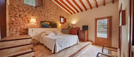 A bedroom with a stone wall, wooden beams, a bed, a desk, and a window.