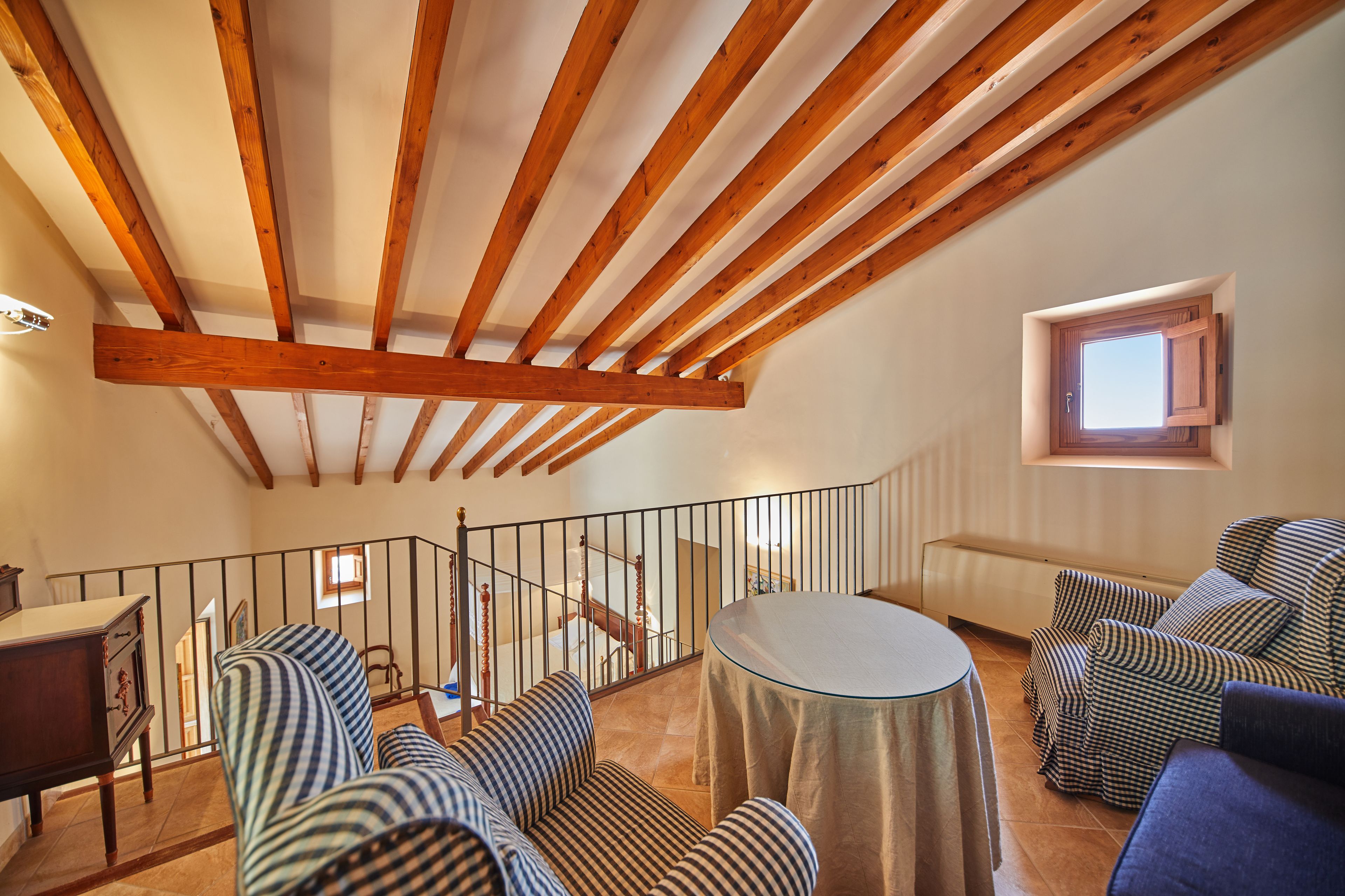 A spacious loft bedroom with exposed wooden beams, a balcony, a round table, and striped sofas.