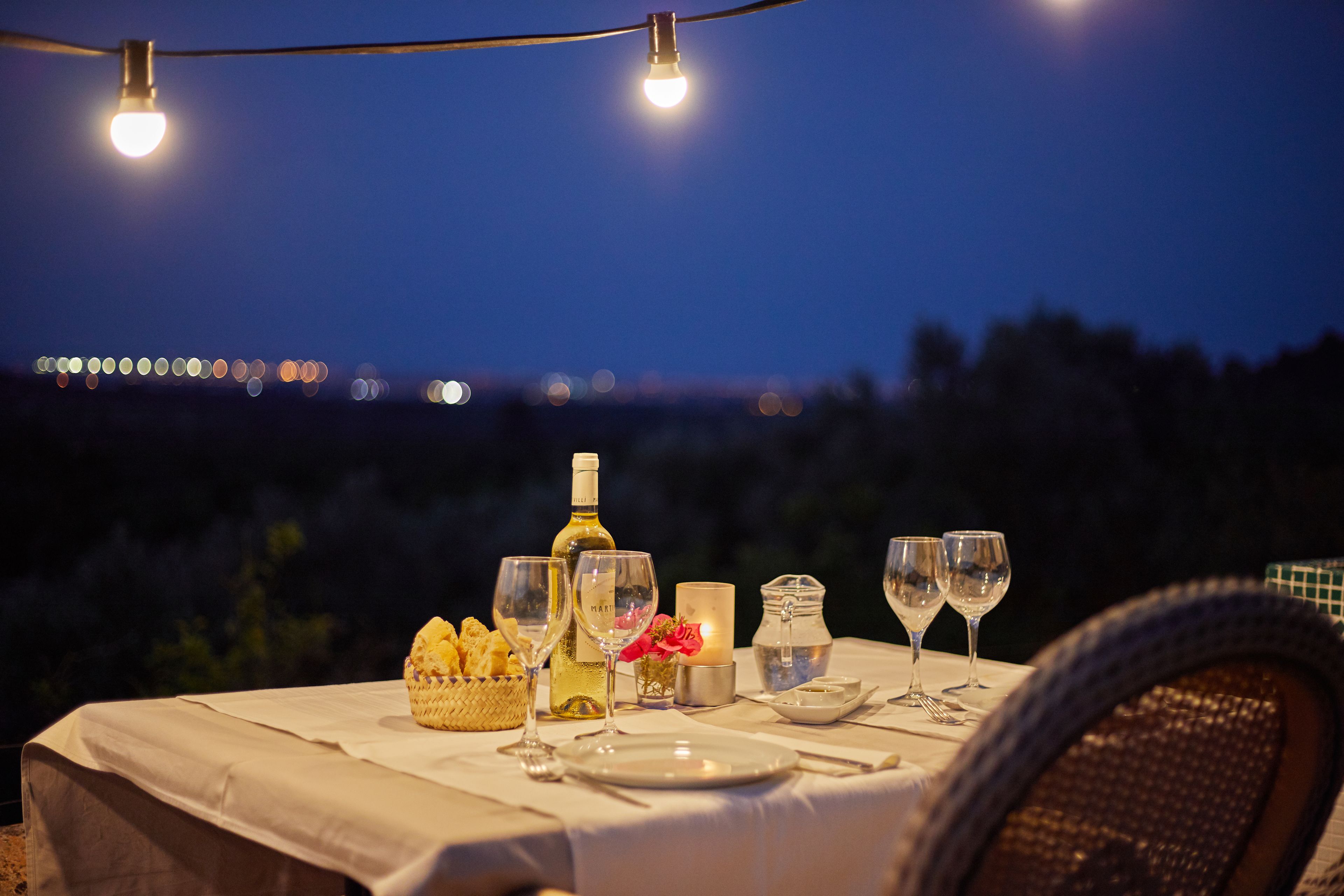 Mesa de comedor al aire libre con platos, vasos, una cesta de pan, una botella de vino y velas, bajo luces colgantes contra un cielo nocturno.
