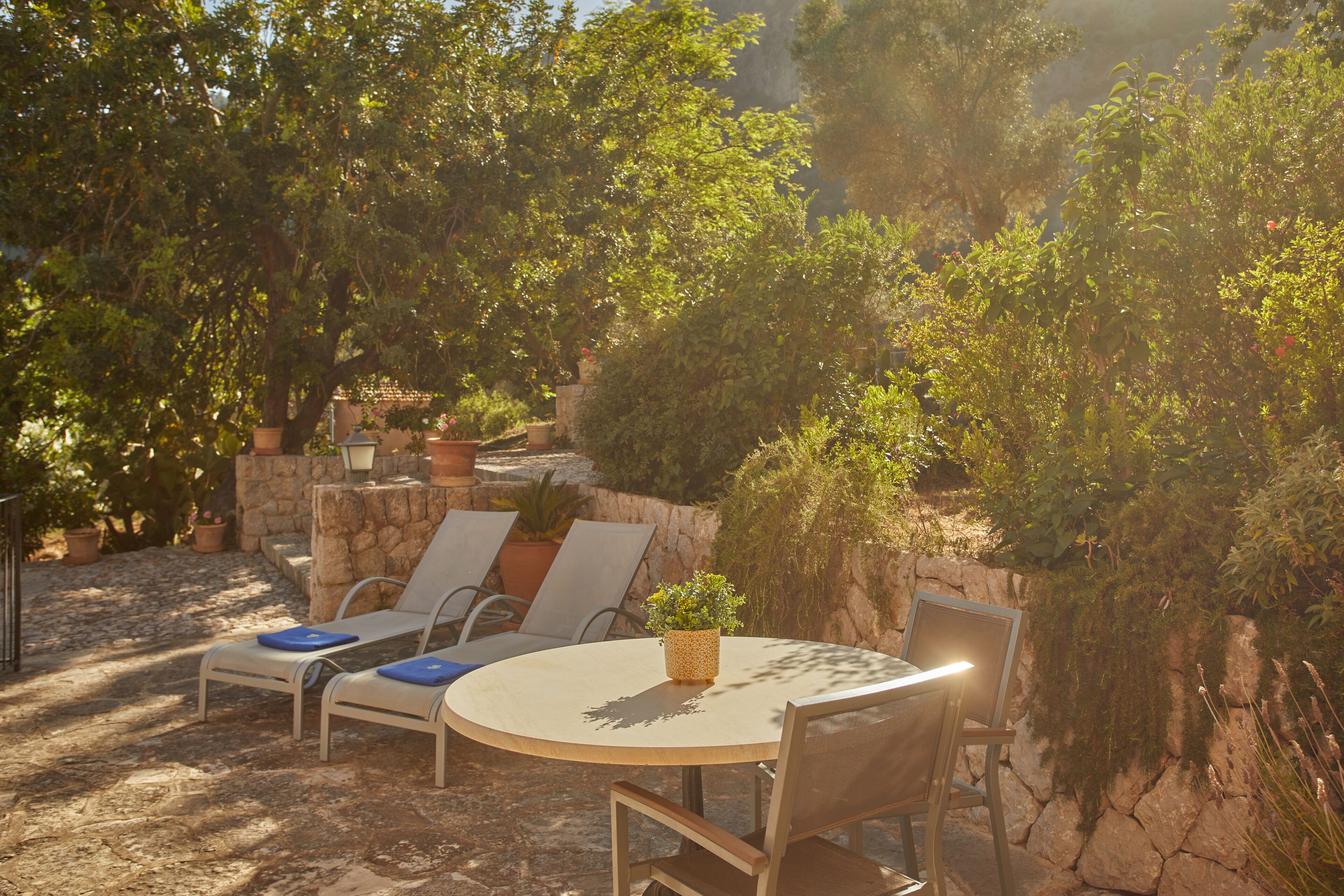 An outdoor seating area with a round table and chairs, surrounded by greenery and stone walls.