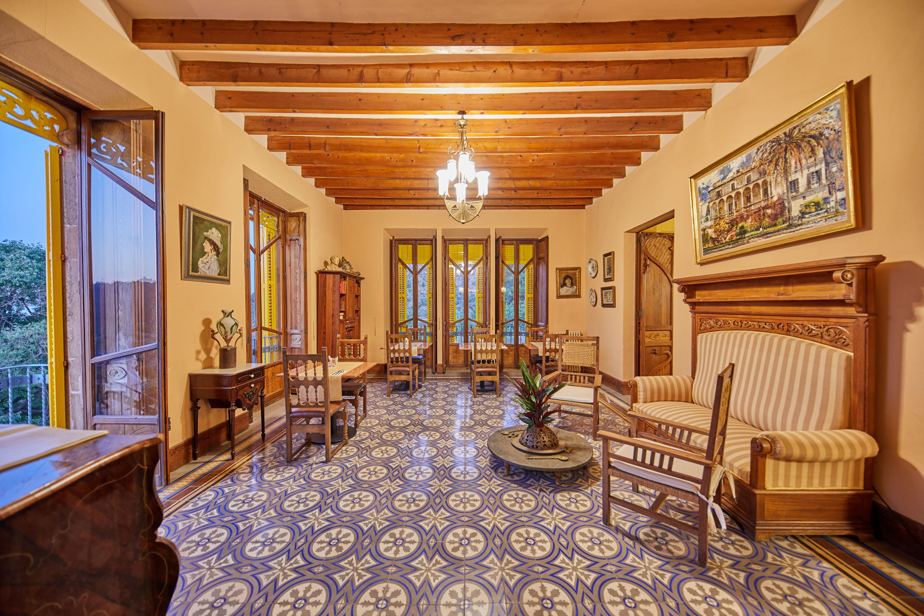 Una habitación amplia con suelo estampado, vigas de madera en el techo y un candelabro. Hay varias sillas y un sofá dispuestos para sentarse.