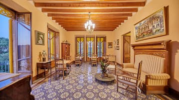 Una habitación amplia con suelo estampado, vigas de madera en el techo y un candelabro. Hay varias sillas y un sofá dispuestos para sentarse.
