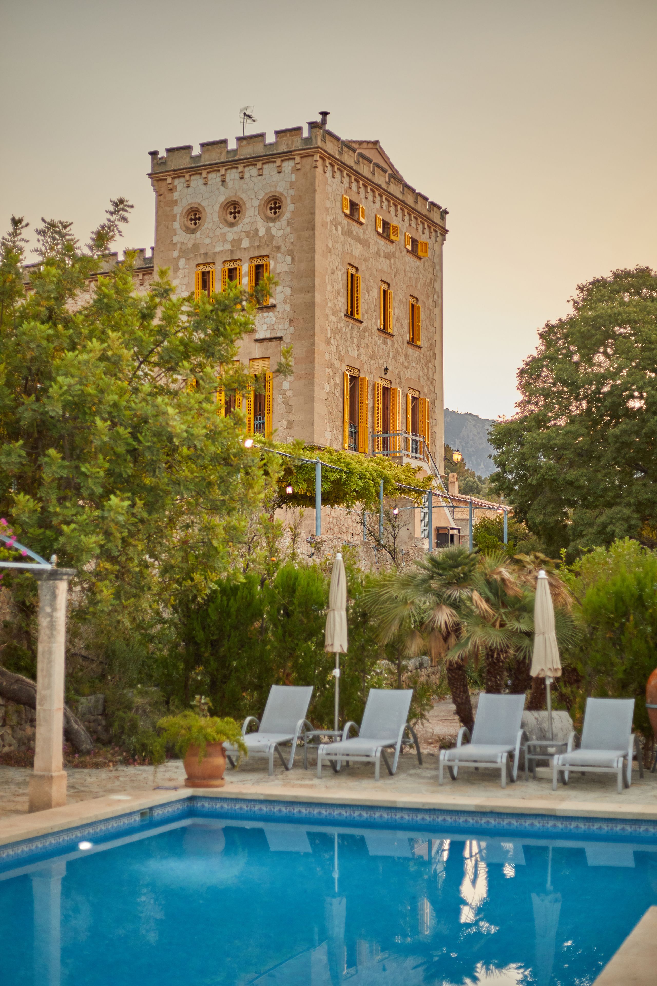 A pool area with lounge chairs and a stone building with yellow windows in the background.
