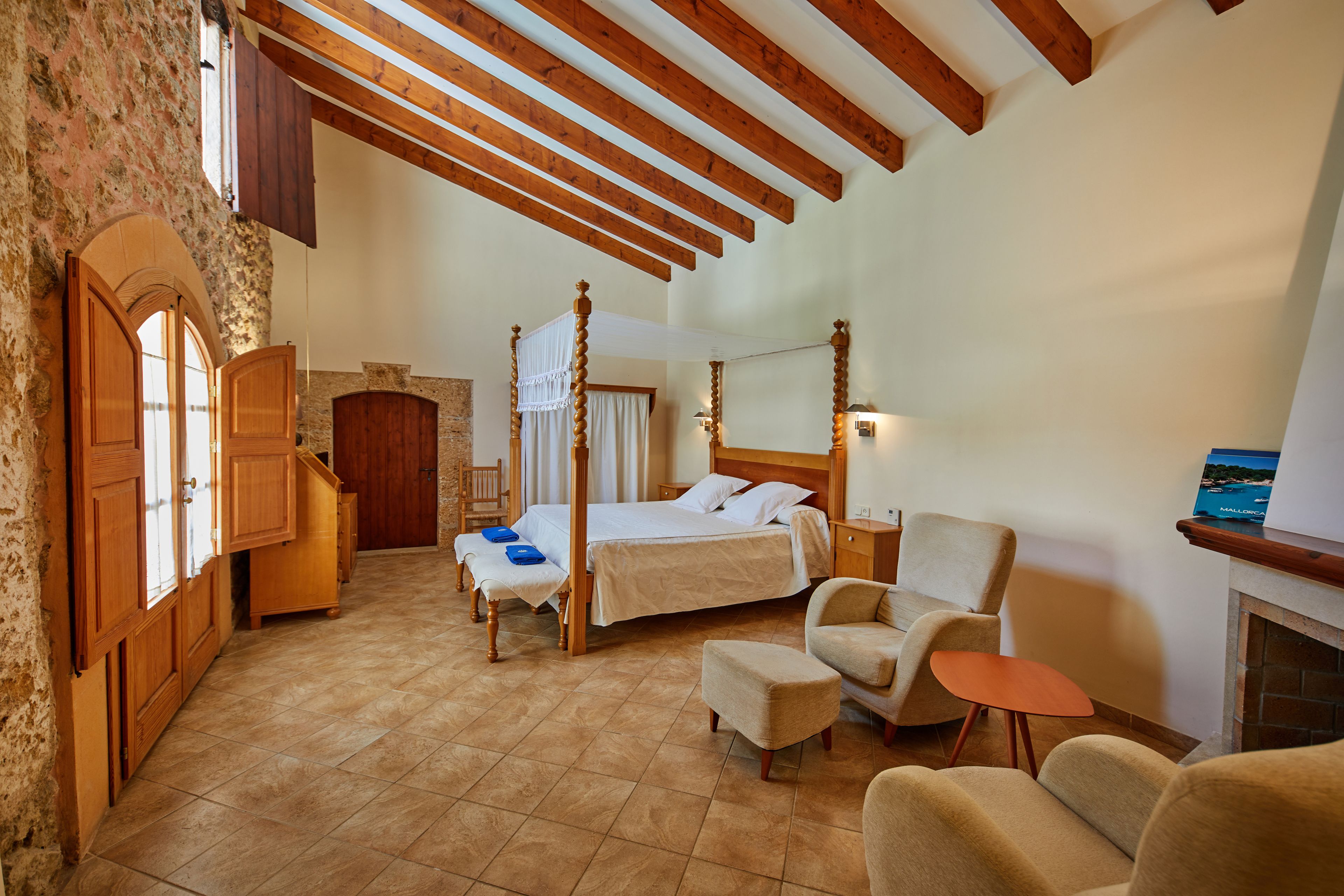 Un dormitorio amplio con una cama grande, un rincón de descanso con una butaca y un reposapiés, y una pared de piedra.
