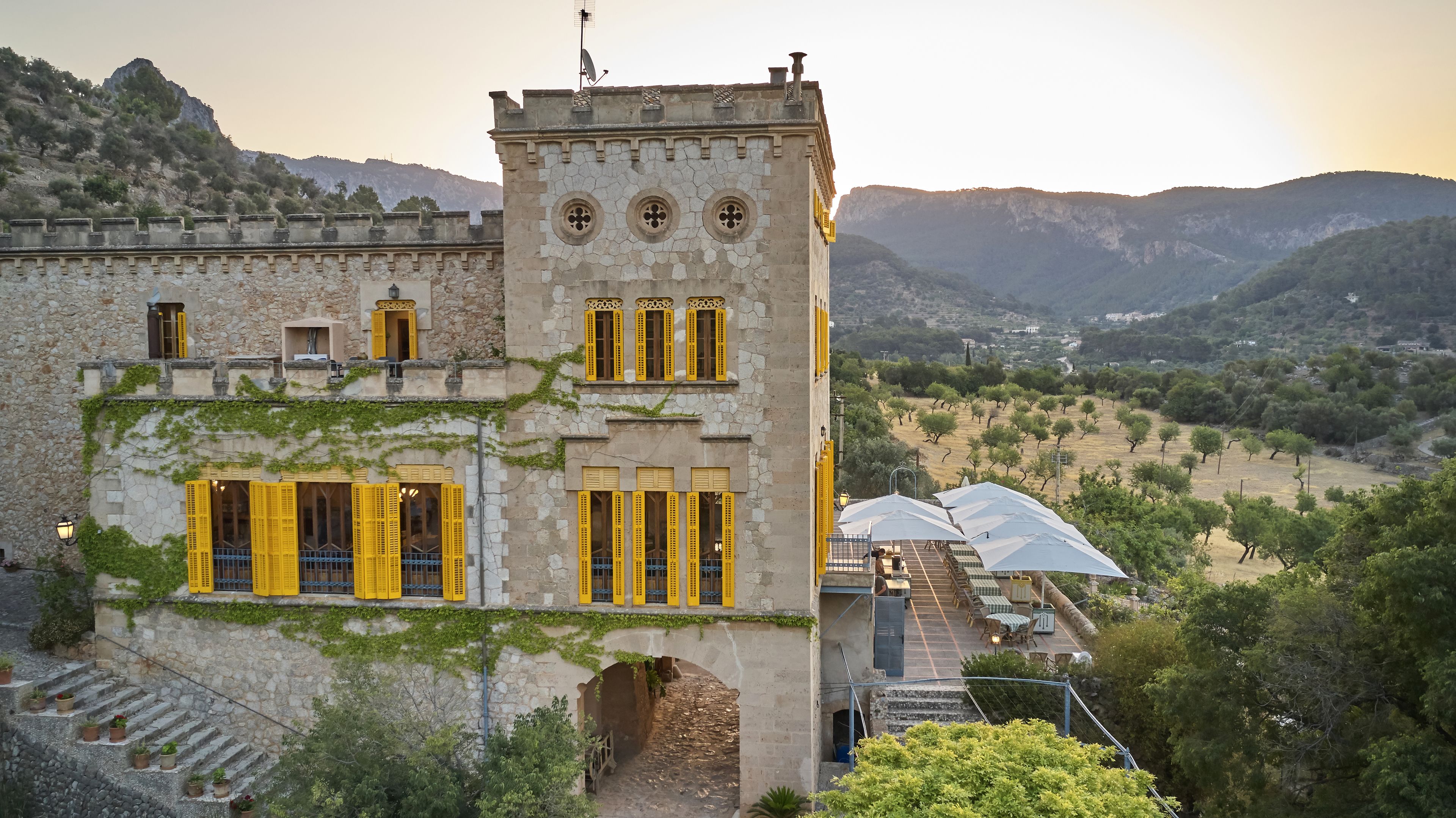 A stone building with yellow shutters, a tower with circular windows, and a view of a valley with trees.