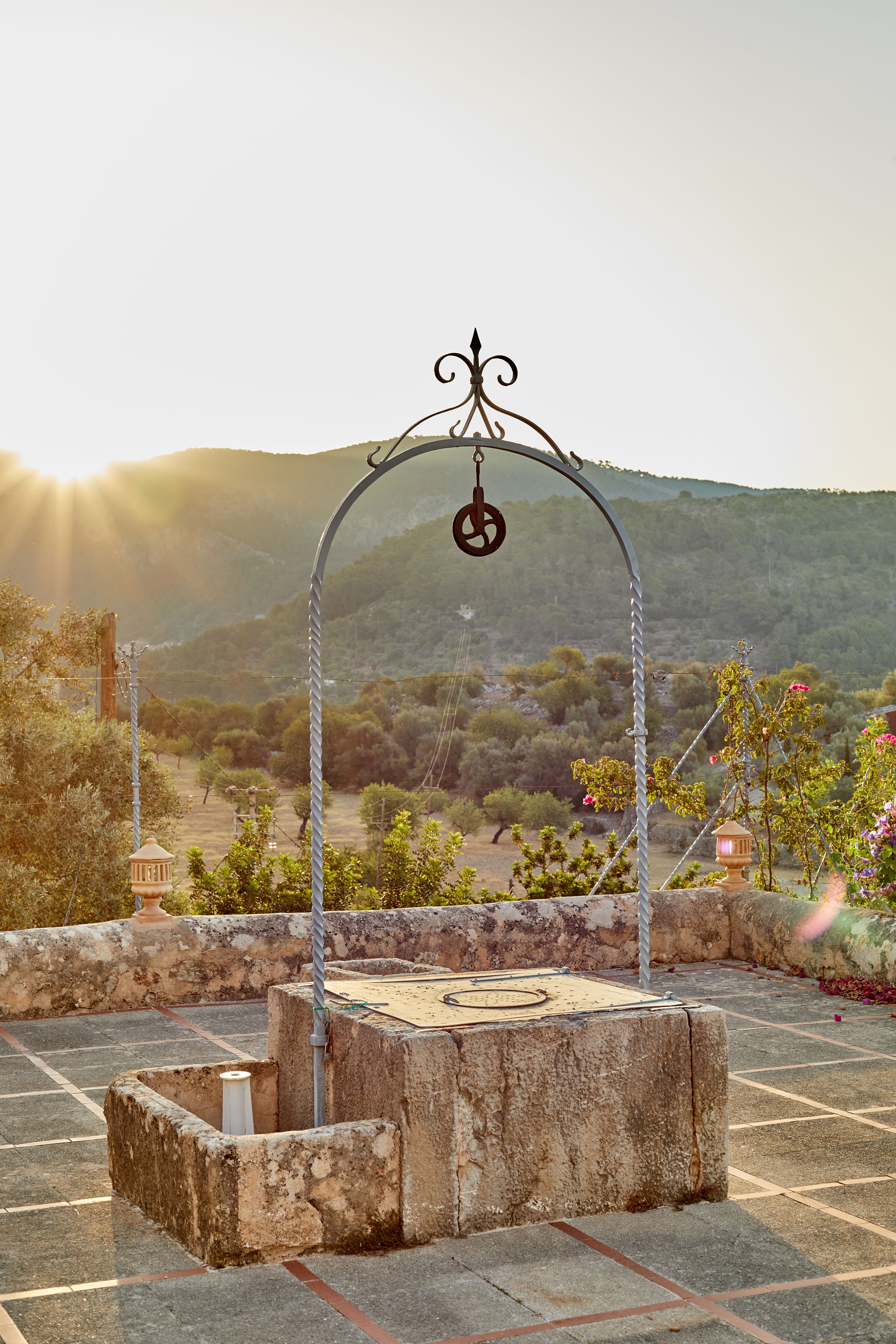 A stone structure with a metal arch and a hanging object, surrounded by a stone wall and greenery, with a mountainous backdrop.
