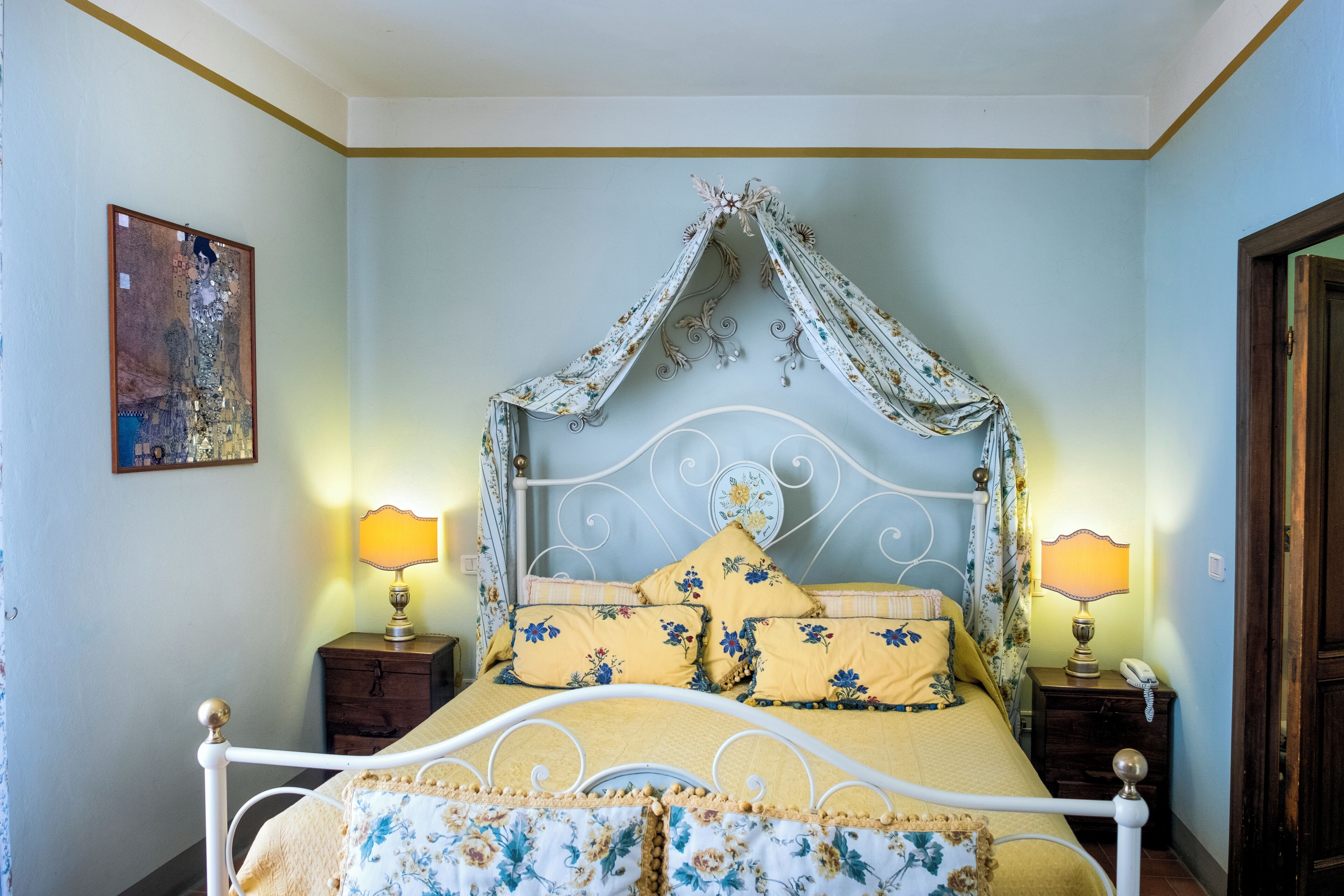 A bedroom with a bed, two bedside tables, a nightstand, a lamp, a wall painting, and a door.
