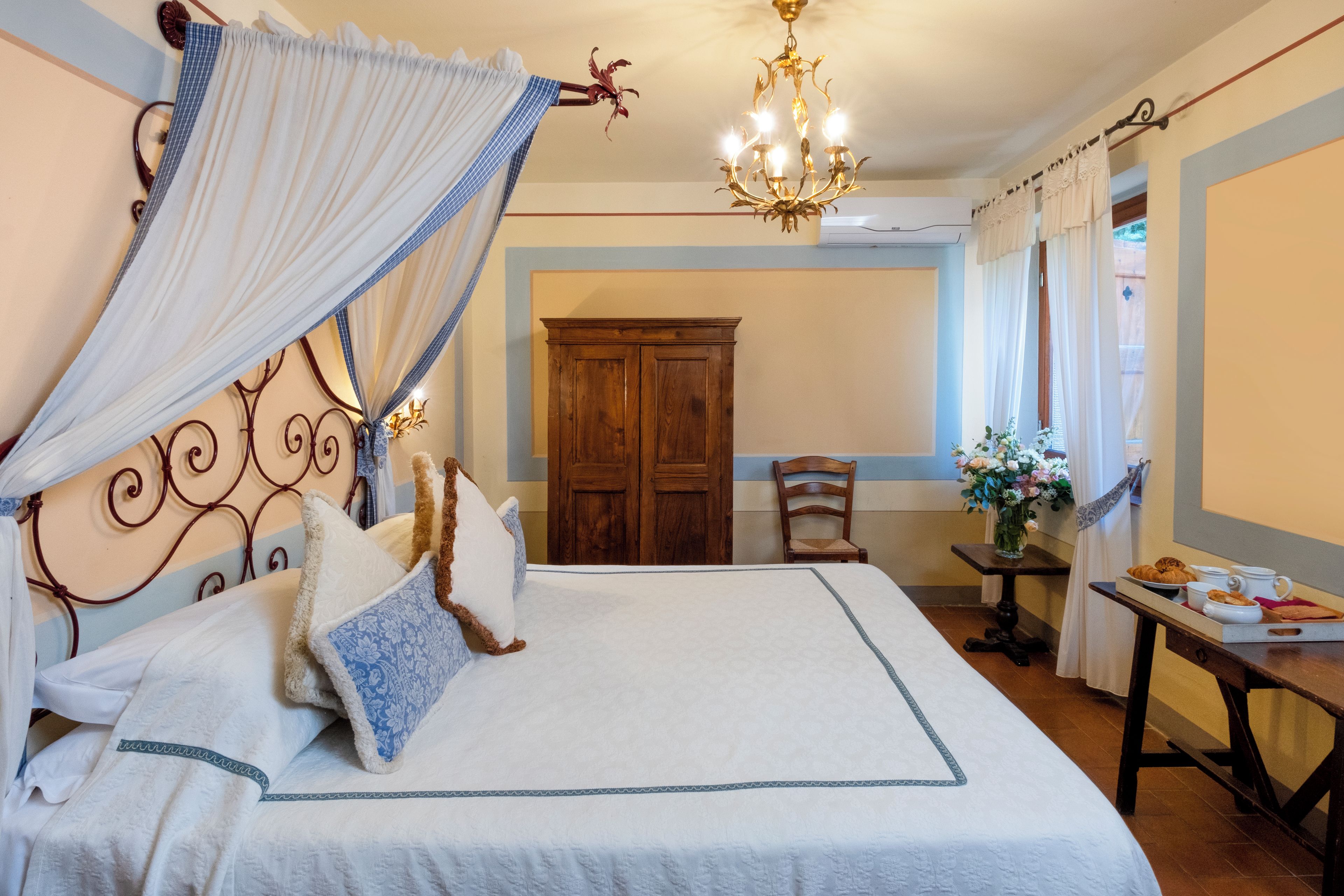 Een net opgemaakt bed met hemel, een houten garderobe, een eettafel met een dienblad eten en een kroonluchter.