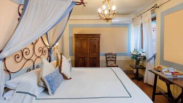Een net opgemaakt bed met hemel, een houten garderobe, een eettafel met een dienblad eten en een kroonluchter.