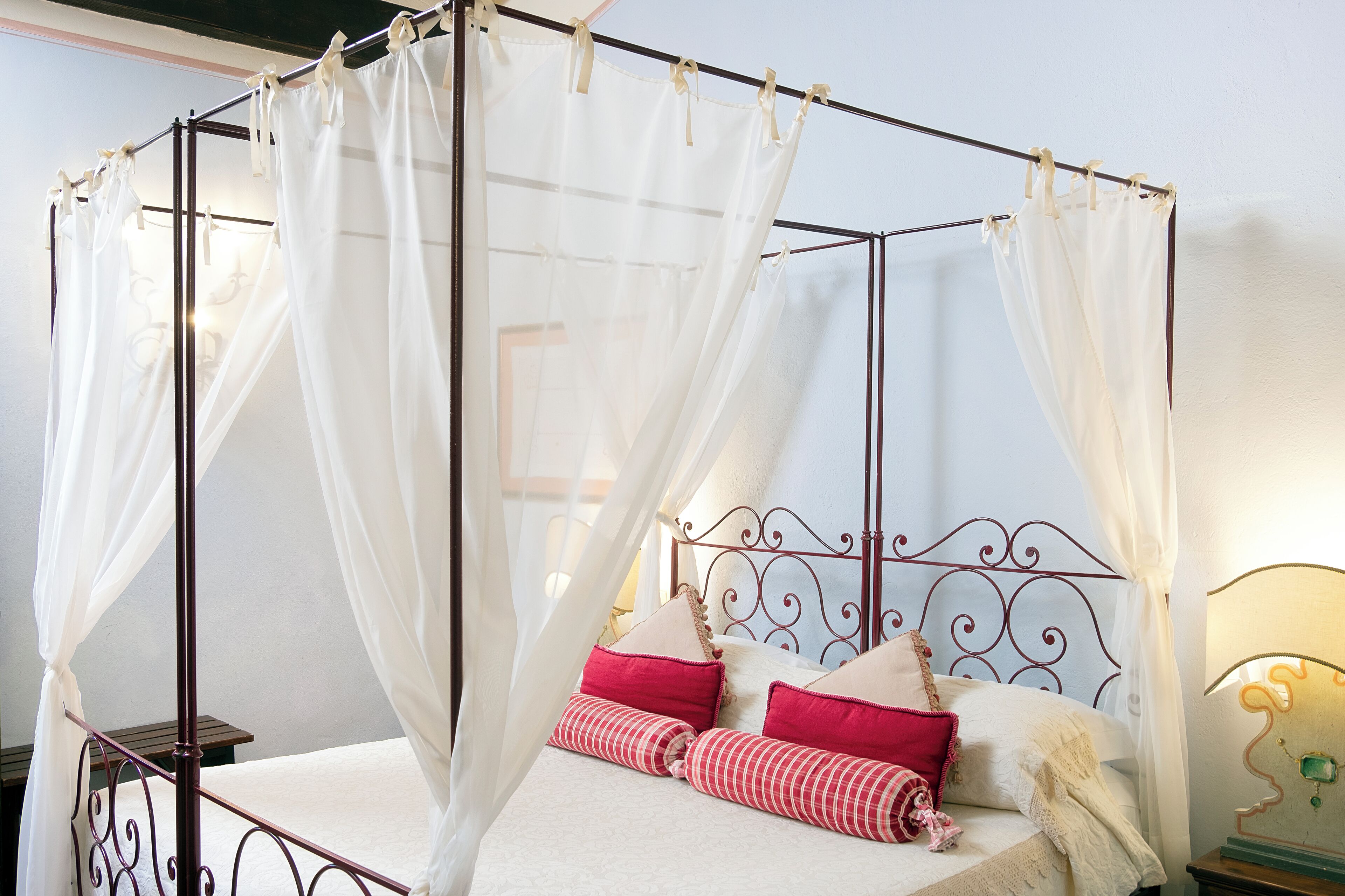Ein Bett mit Baldachin, weiße Vorhänge, rote und weiße Kissen sowie ein Metall-Kopfstück.