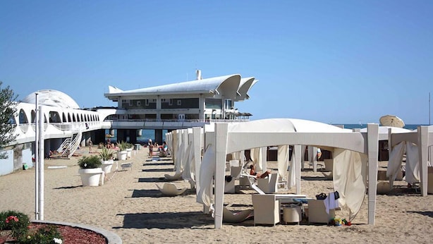 A beachside hotel with outdoor seating areas, a large building with a unique roof design, and a clear view of the sea.