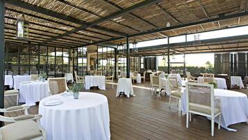 Un área de comedor al aire libre con mesas redondas cubiertas de manteles blancos, sillas de mimbre e iluminación colgante.