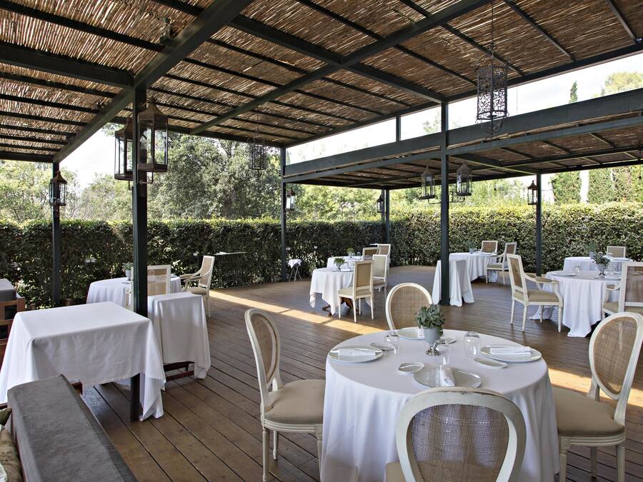 Un patio al aire libre con mesas y sillas redondas, cubierto por un techo de madera, rodeado de follaje.
