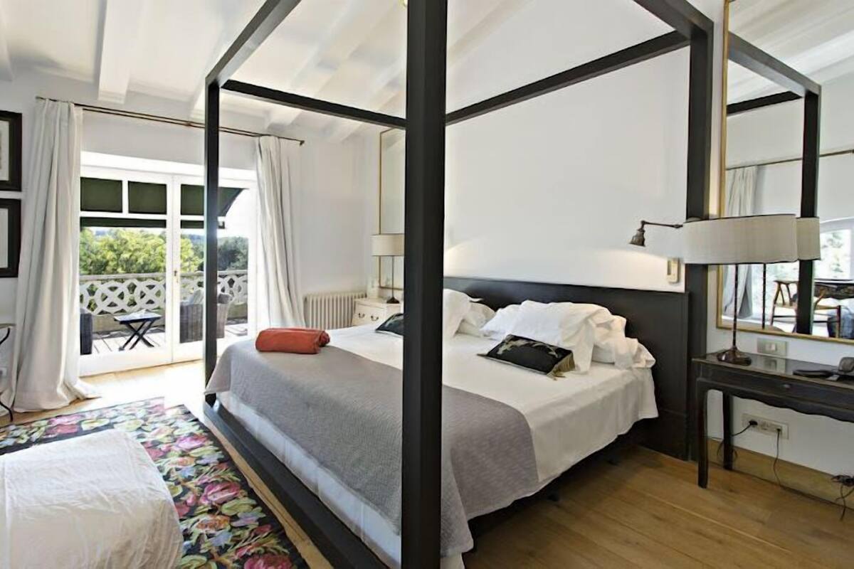 A four-poster bed with a grey and white bedspread, a bedside table with a lamp, and a large mirror on the wall.