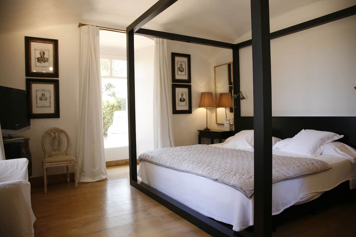 A bedroom with a four-poster bed, a chair, a desk with a lamp, and framed pictures on the wall.