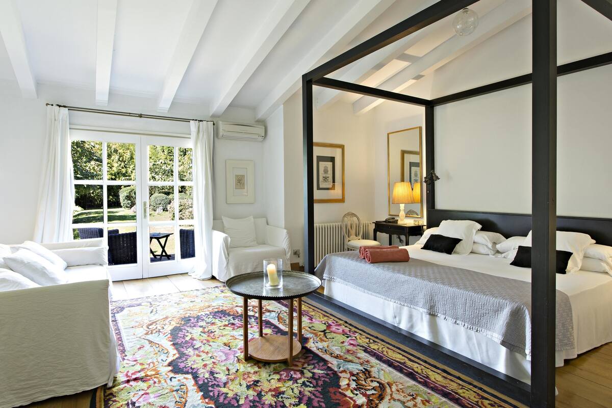 A bedroom with a large bed, a small round table, a chair, and a colorful rug.