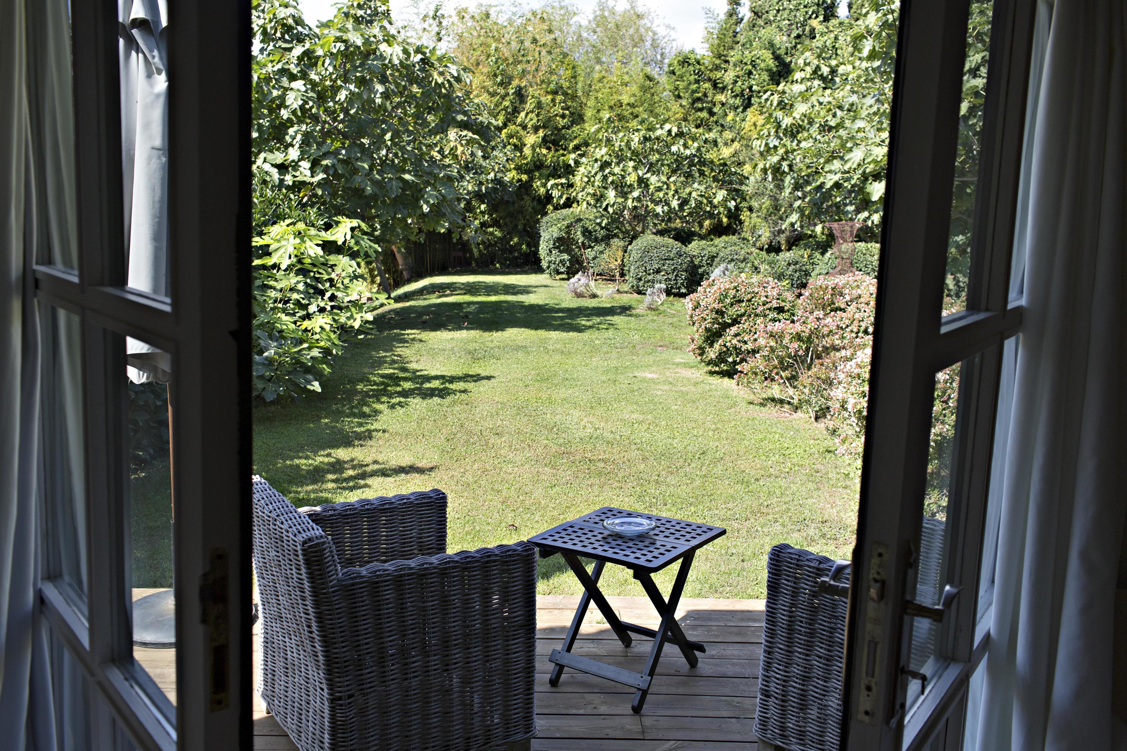 An open door to a patio with wicker furniture and a small table, overlooking a well-maintained garden with various plants and a lawn.