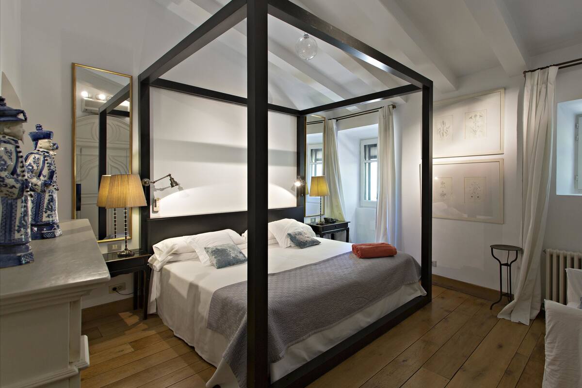 A four-poster canopy bed with white linens and a grey throw, a bedside table with a lamp, and a nightstand with a phone.