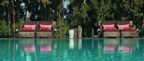 Zona junto a la piscina con sillones de mimbre y cojines rojos, rodeada de árboles.