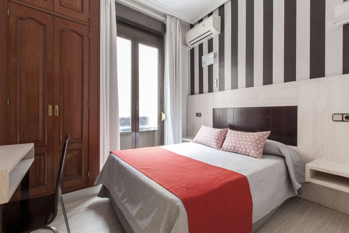 A hotel room with a large bed, a wooden wardrobe, a desk, and a window with curtains.