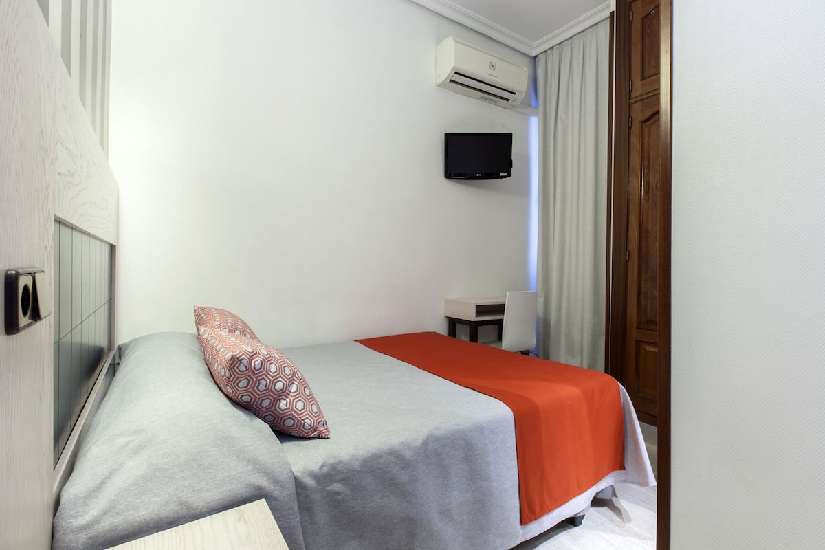 A single bed with a grey and orange bedspread, a patterned pillow, a mounted television, and a wooden door.