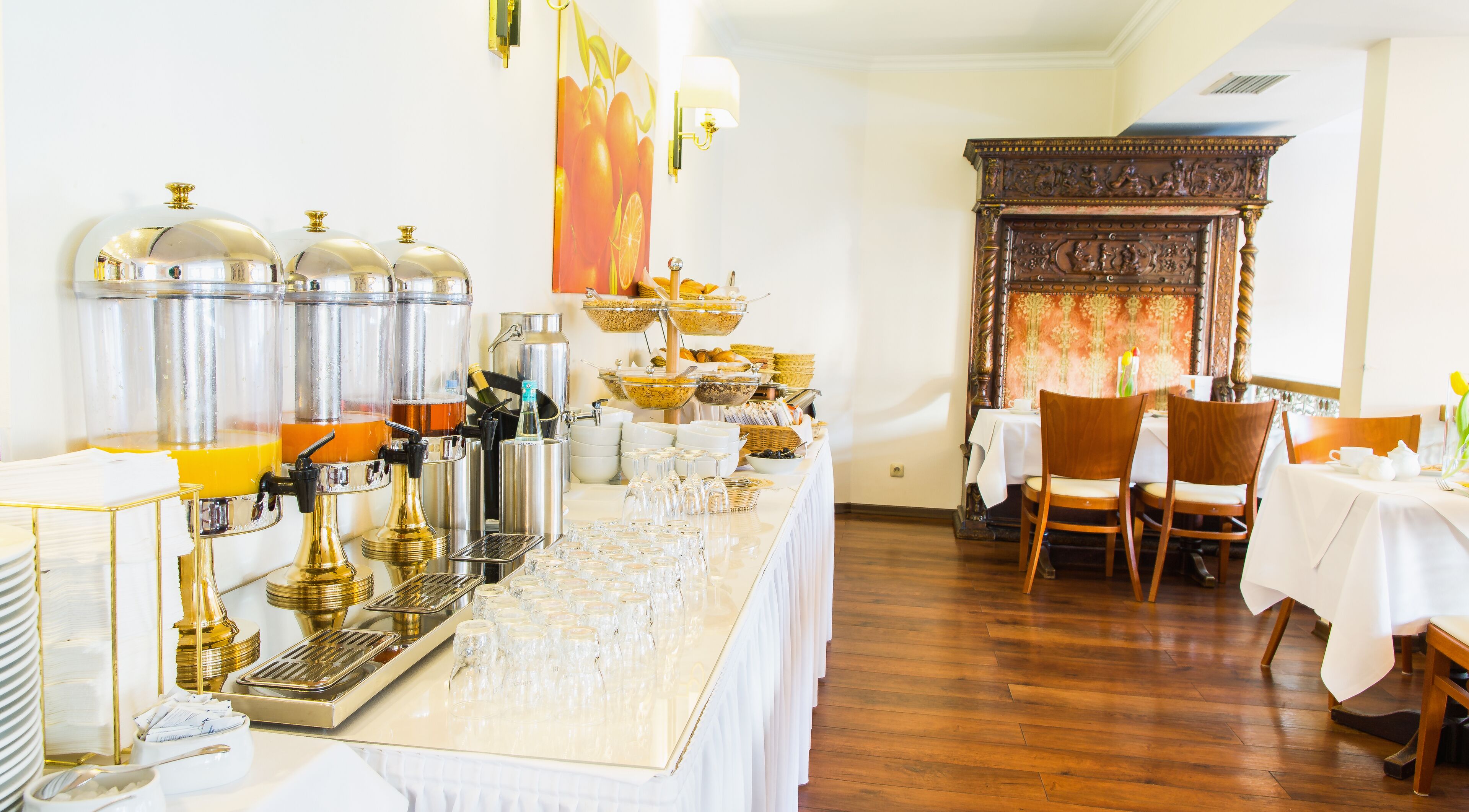 Disposição de buffet com diversos dispensadores de comida e bebida, área de refeições com mesas e cadeiras, e parede decorativa com padrão floral.