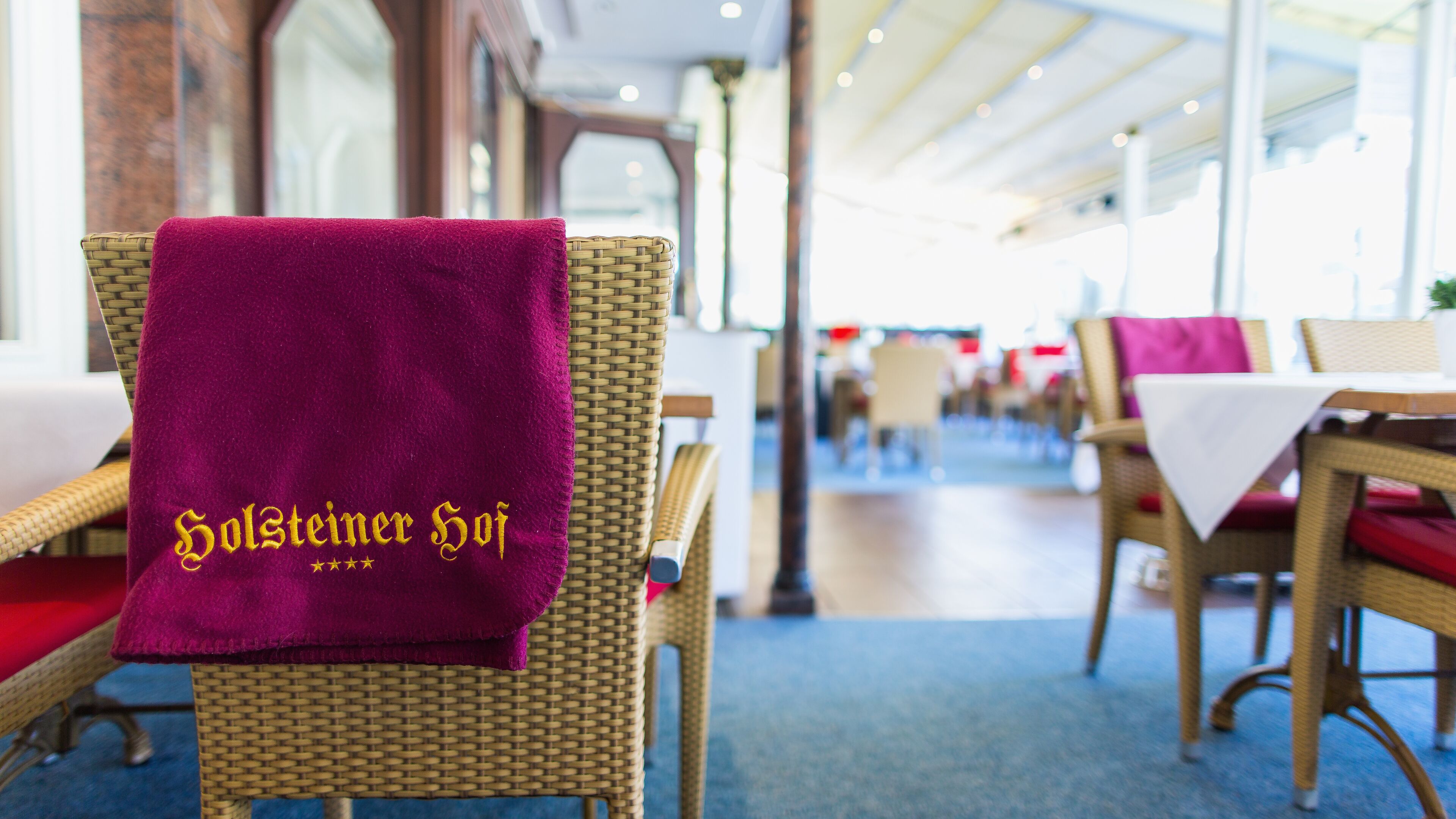 Ein Essbereich mit Korbstühlen und Tischen, einige mit roten Kissen. Ein lilafarbener Handtuch mit der Aufschrift "Holsteiner Hof" ist über einen Stuhl gelegt.
