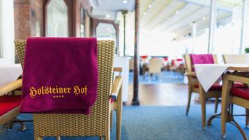 Ein Essbereich mit Korbstühlen und Tischen, einige mit roten Kissen. Ein lilafarbener Handtuch mit der Aufschrift "Holsteiner Hof" ist über einen Stuhl gelegt.