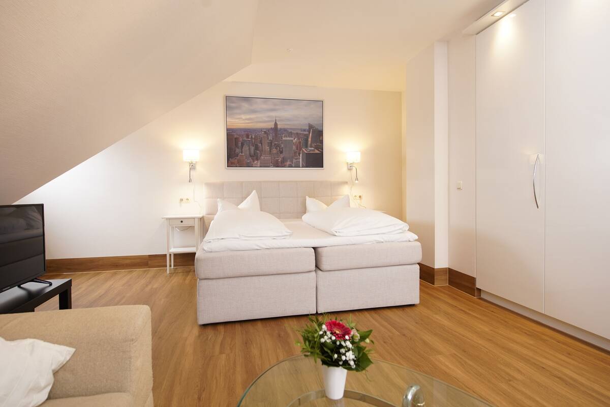 A modern hotel room with a bed, a television, a glass table with a flower vase, and a cityscape painting on the wall.