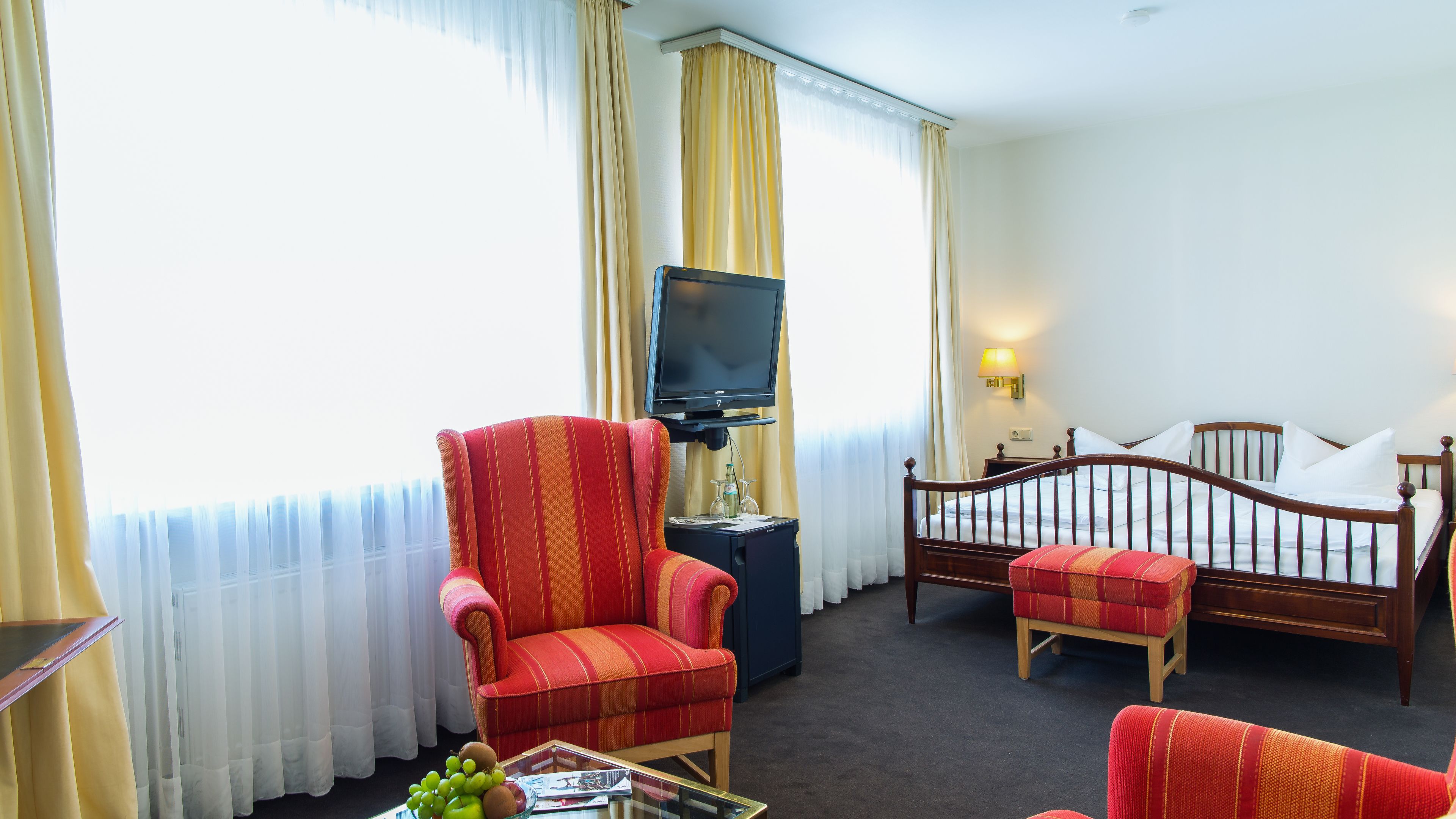 Habitación de hotel con una cama, un televisor en un soporte, un sillón rojo, una silla de madera y una pequeña mesa con frutas y una revista.