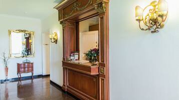 A hallway with a wooden reception desk, a mirror, a vase with flowers, and wall sconces.