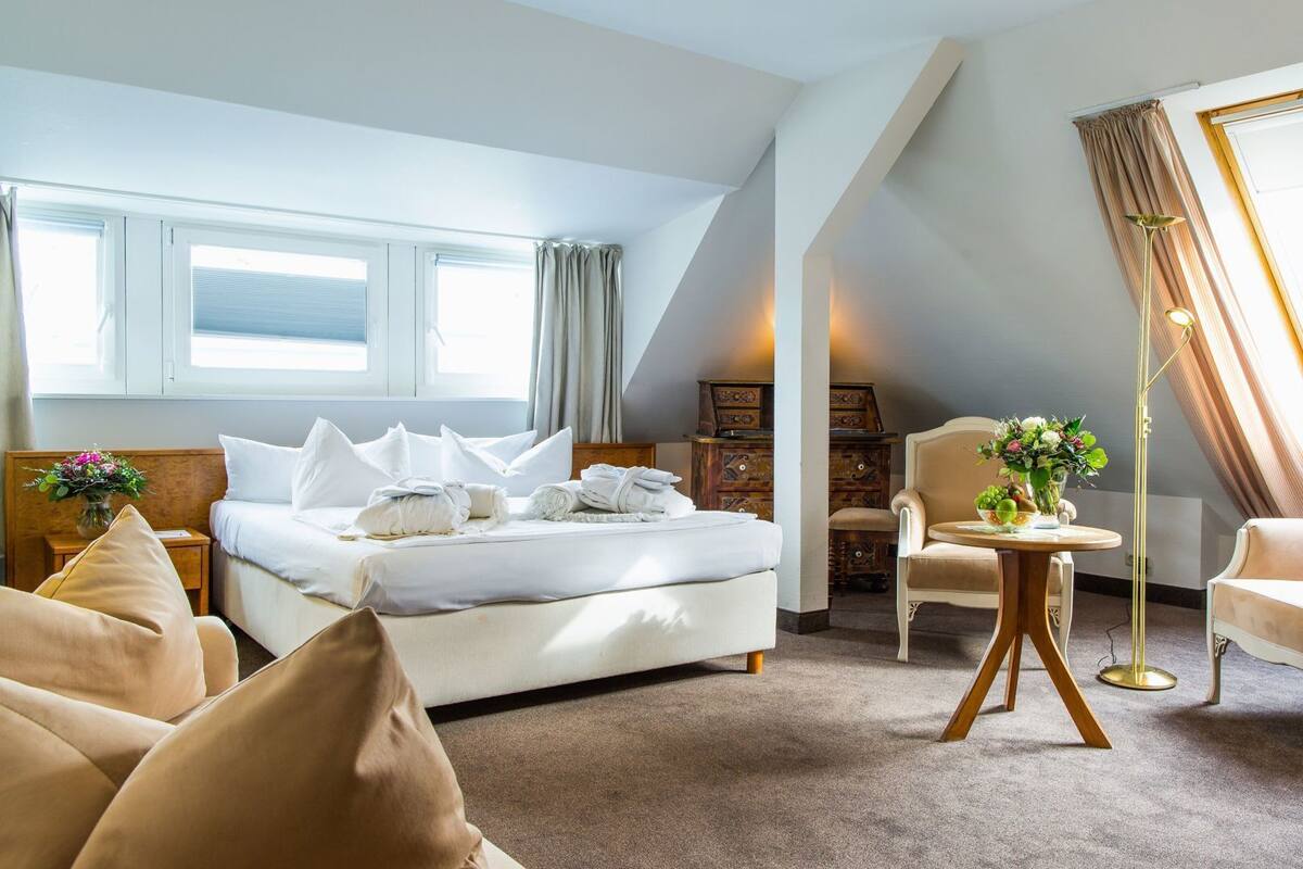 A hotel room with a bed, two armchairs, a small table, a desk, and a window with curtains.